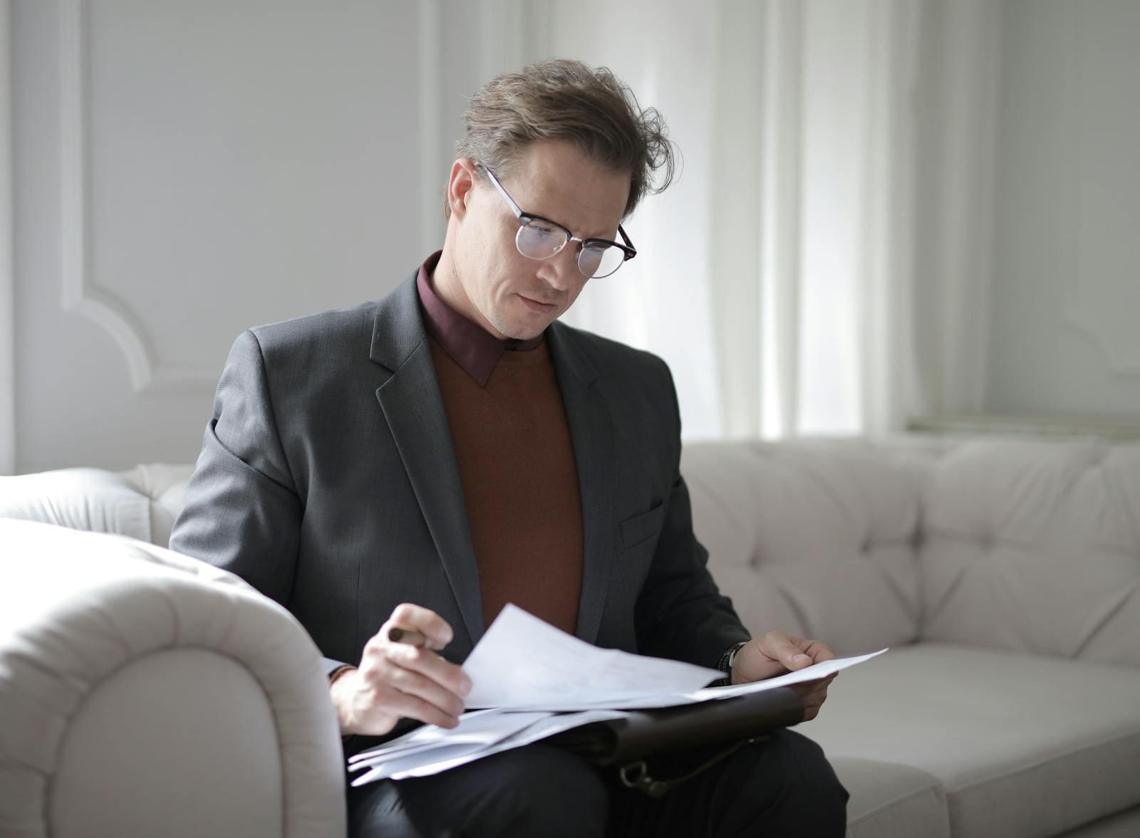 A man in a jacket reading through a sheaf of papers on a sofa