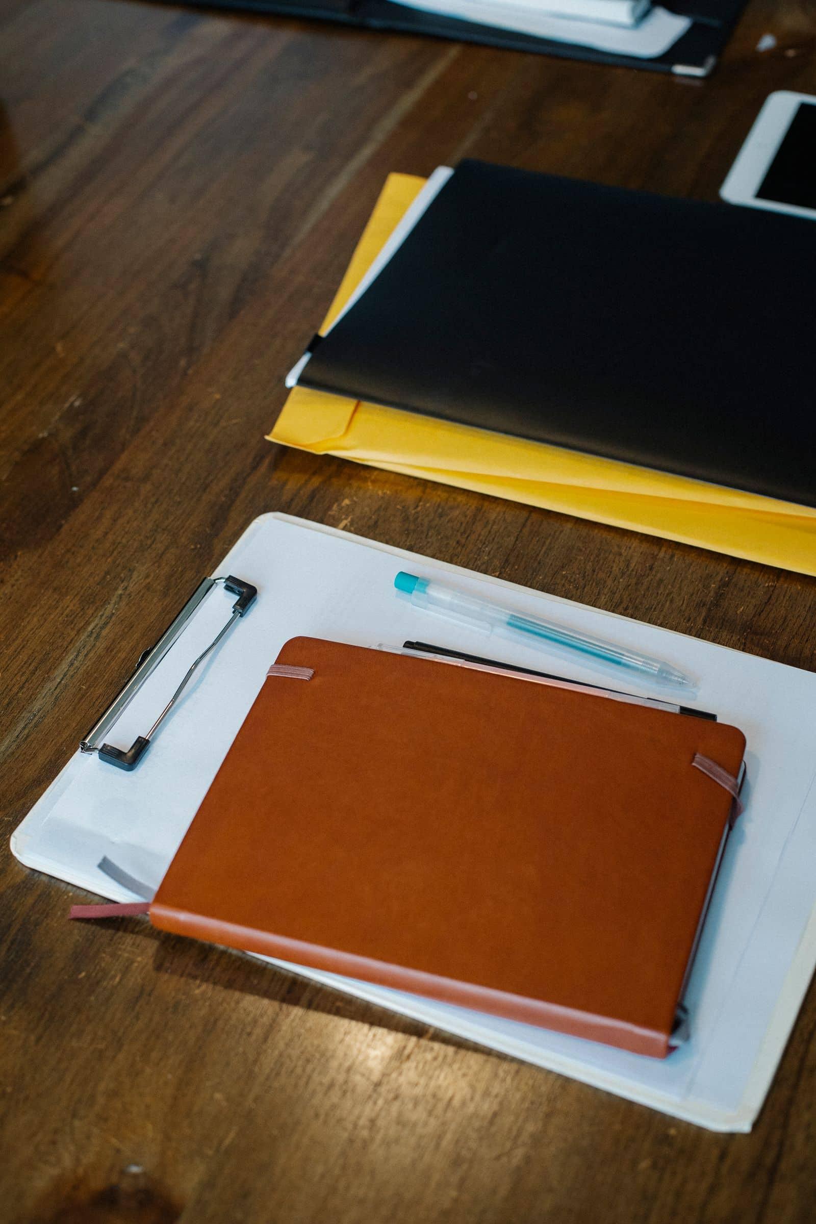 A clipboard of blank forms, folders and pens laid out on a wooden desk