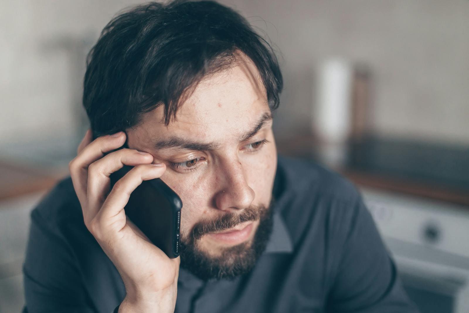 A bearded man on a phone call indoors, looking down as he listens